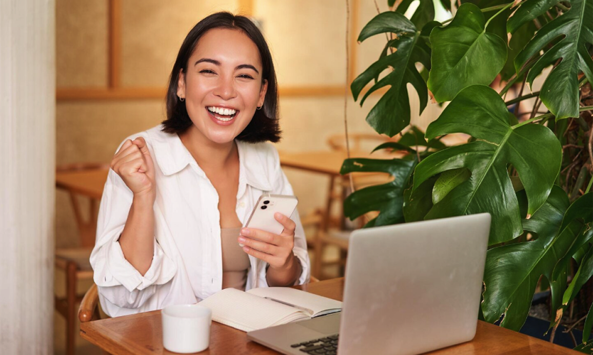 Smiling Wix merchant celebrating with phone and laptop at a sunny desk with a plant